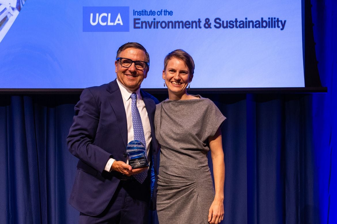 Tony Pritzker (left), in a navy suit, and Zuzana Buřivalová, in a gray dress, smile while holding an award onstage beneath a UCLA Institute of the Environment & Sustainability sign.