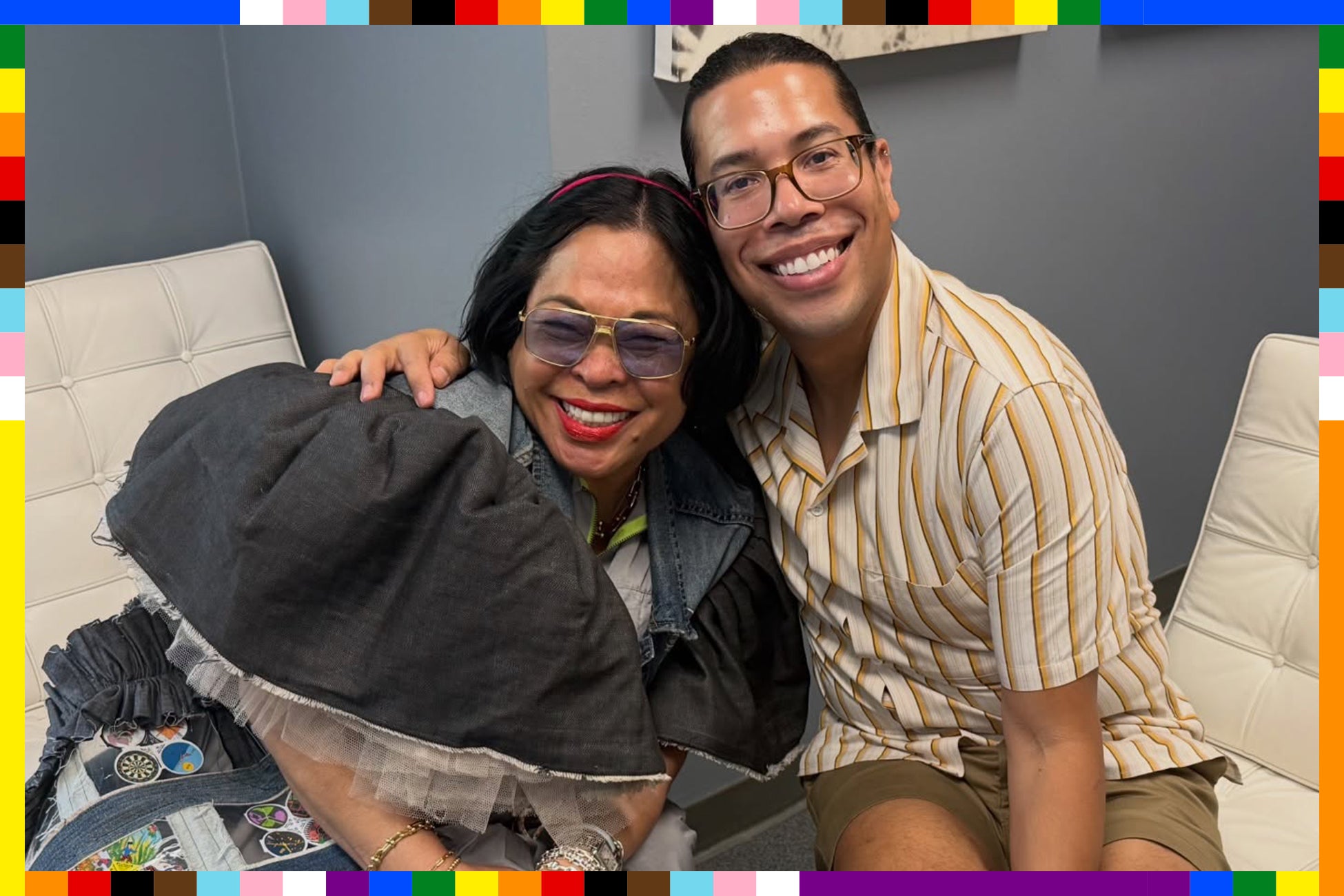 A woman and man sit on a couch smiling. Vibrant rainbow-striped outline surrounds image.