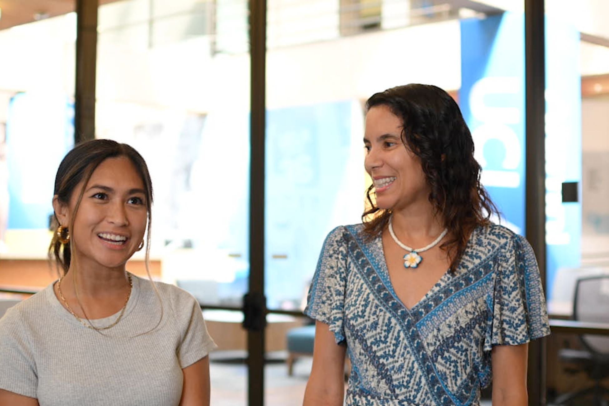 Mentor and Mentee smiling and chatting in a bright room in the James West Alumni Center, with large windows and computer workstations in the background.
