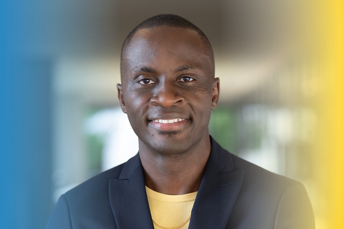 Idriss Njike smiles at the camera in a close-up portrait, wearing a dark blue blazer and yellow shirt against a softly blurred background accented with UCLA blue and gold.