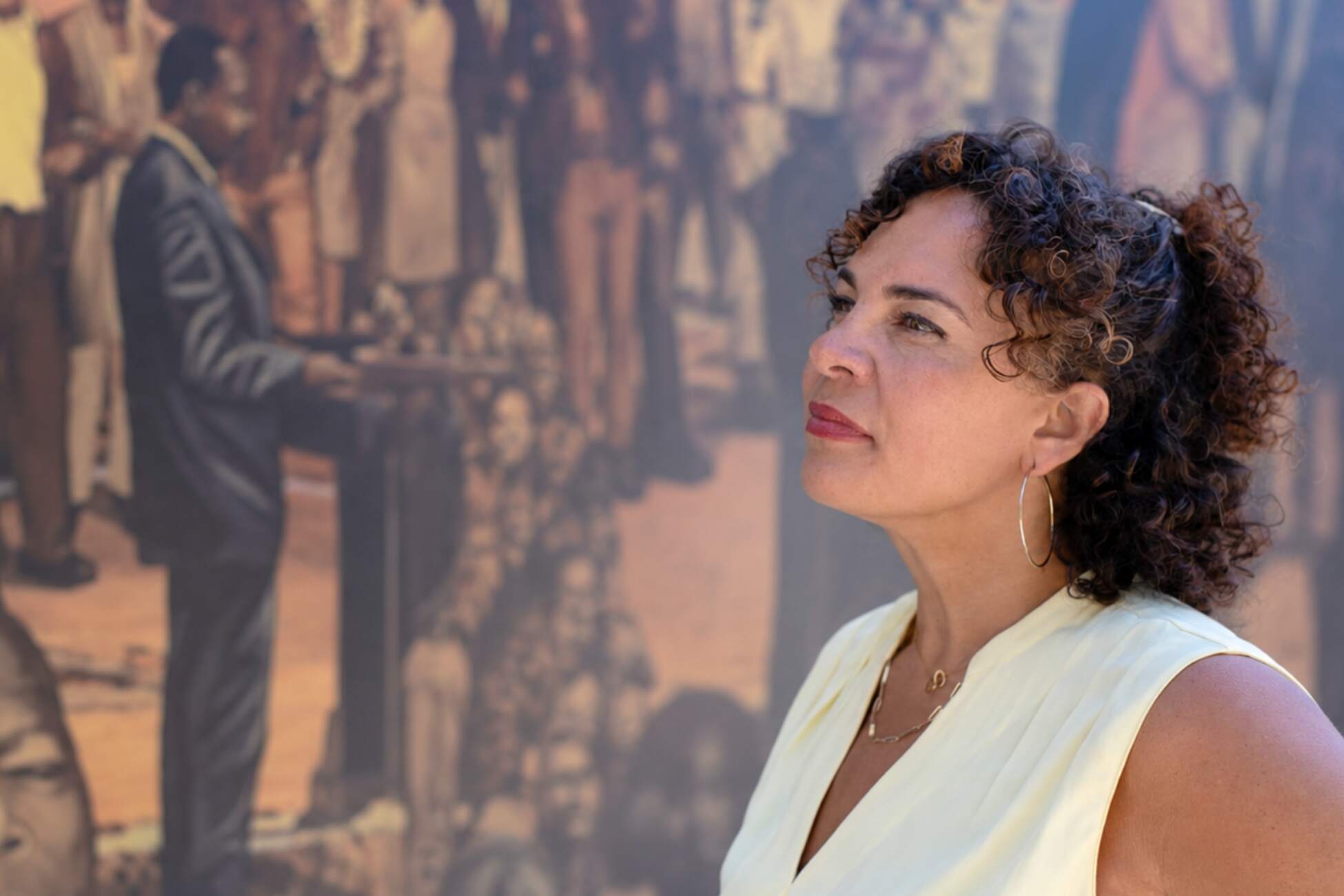 A woman with curly hair and hoop earrings thoughtfully gazes at a mural depicting civil rights activists, leaders, and prominent individuals. 