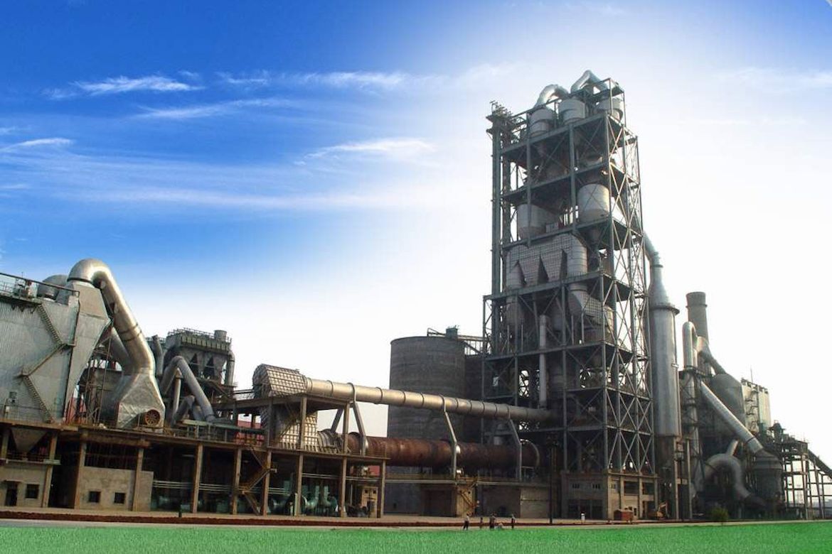 Industrial cement production facility with a rotary kiln, clinker-processing equipment, large ducts, towers and interconnected machinery beneath a blue sky.