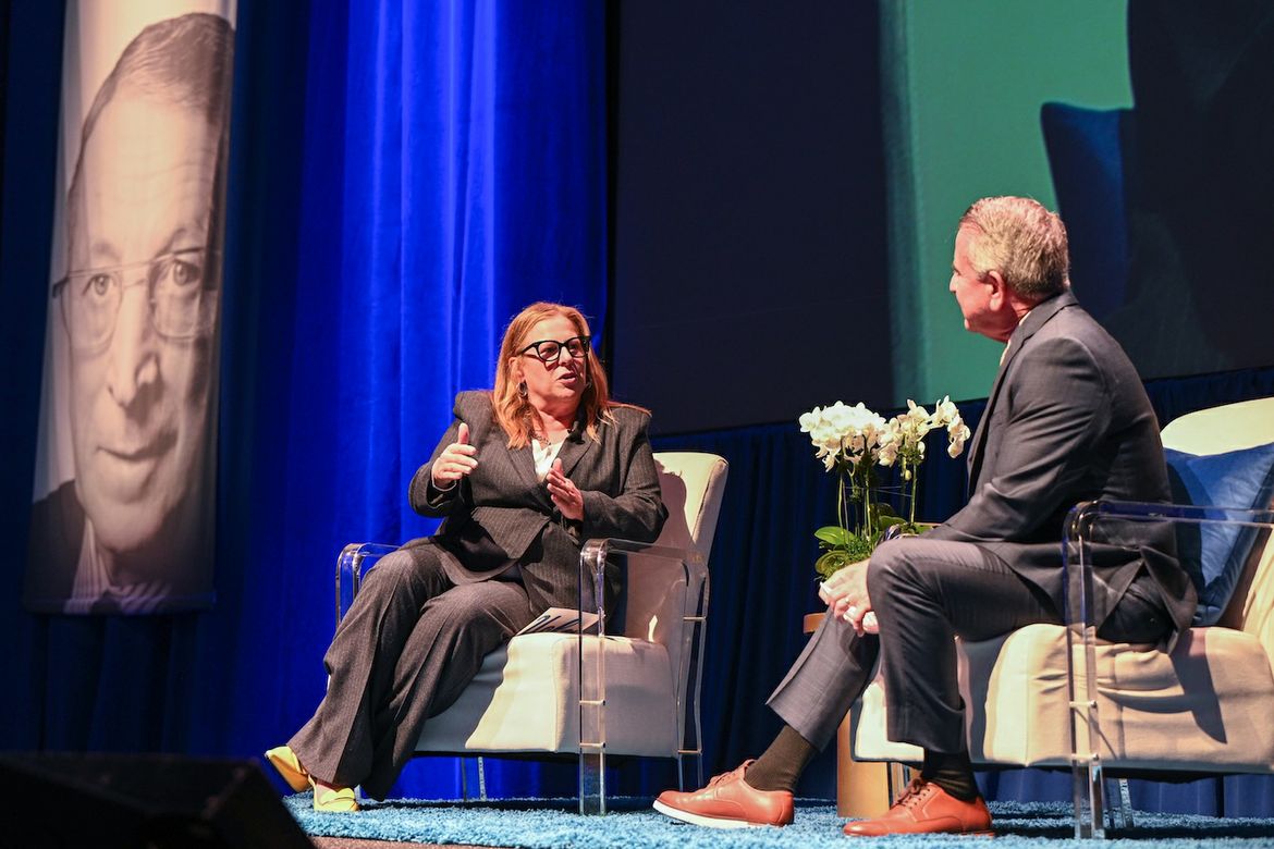 Cori Close gestures while speaking with Brian Cornell onstage at Pauley Pavilion, with a large portrait of John Wooden visible behind them.
