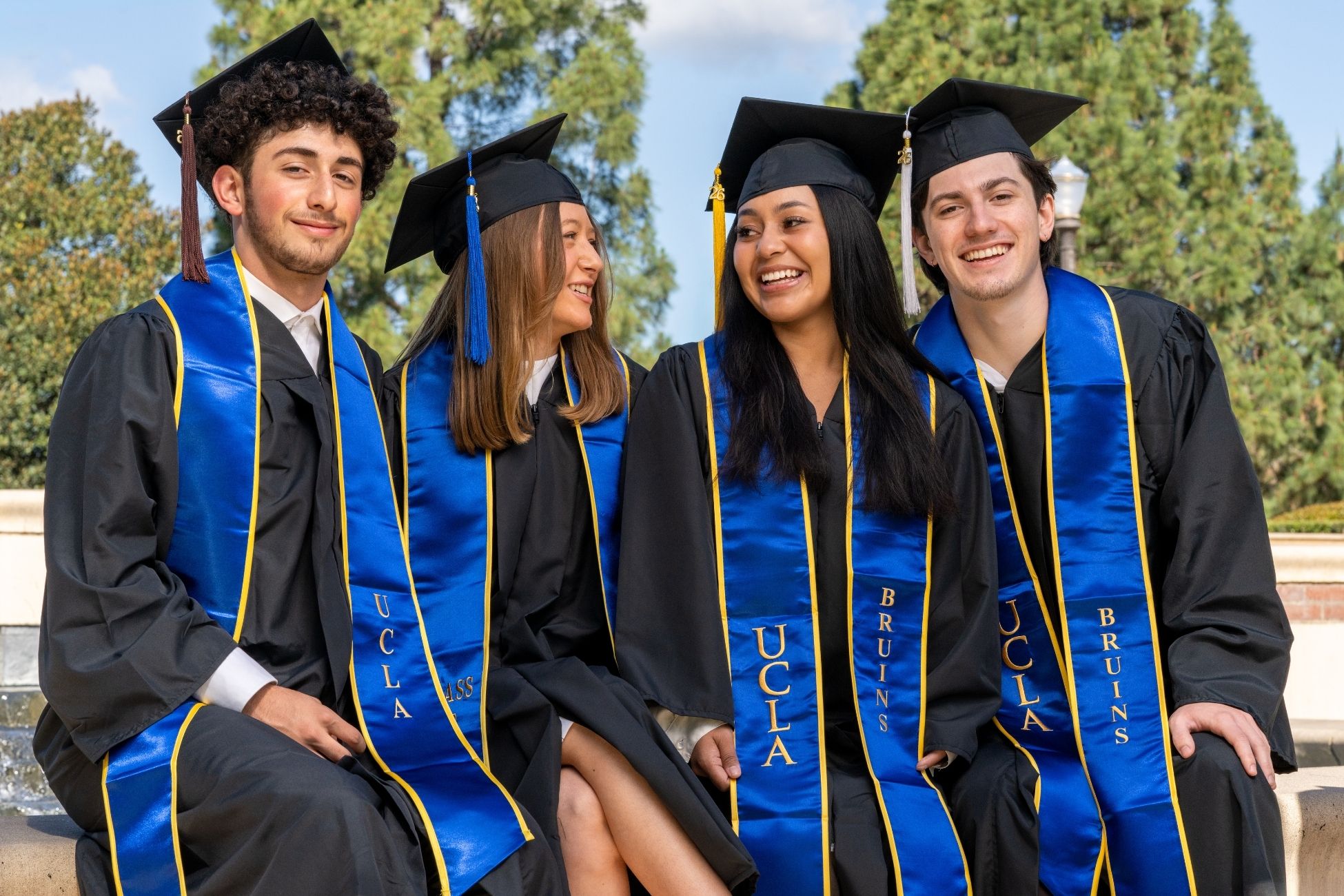 Four graduates wearing black gowns, caps, and blue stoles, smile and look towards each other.