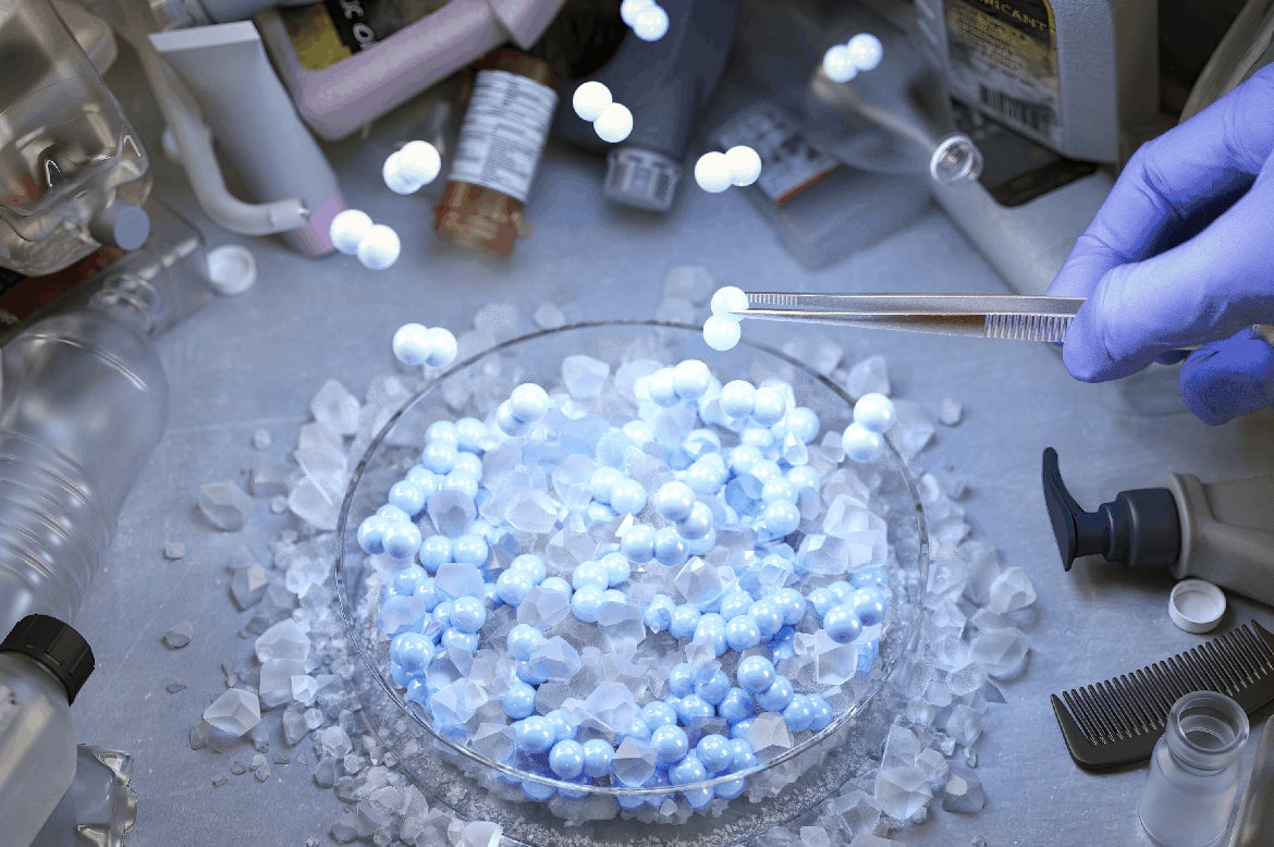 Illustration showing a gloved hand using tweezers to hold a glowing blue bead above a dish filled with similar beads and translucent crystals, surrounded by laboratory tools and plastic containers.
