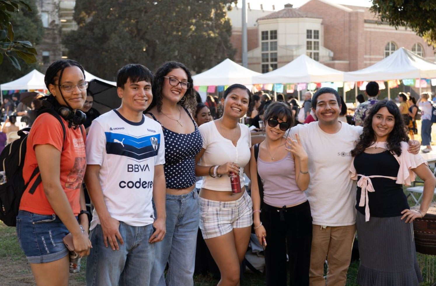 A group of eight UCLA students pose together at an outdoor campus fair with white vendor tents and colorful decorations in the background. A brick university building and trees are visible behind them.