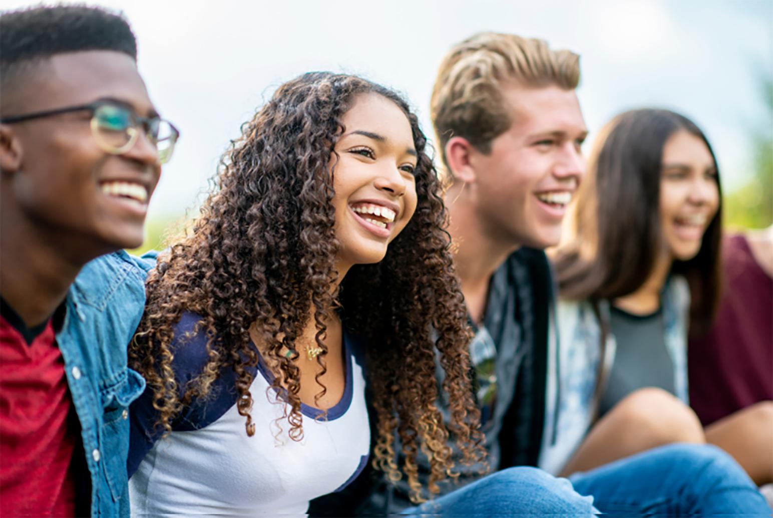 A diverse group of five young adults sitting outdoors, smiling and laughing together in a relaxed social setting.