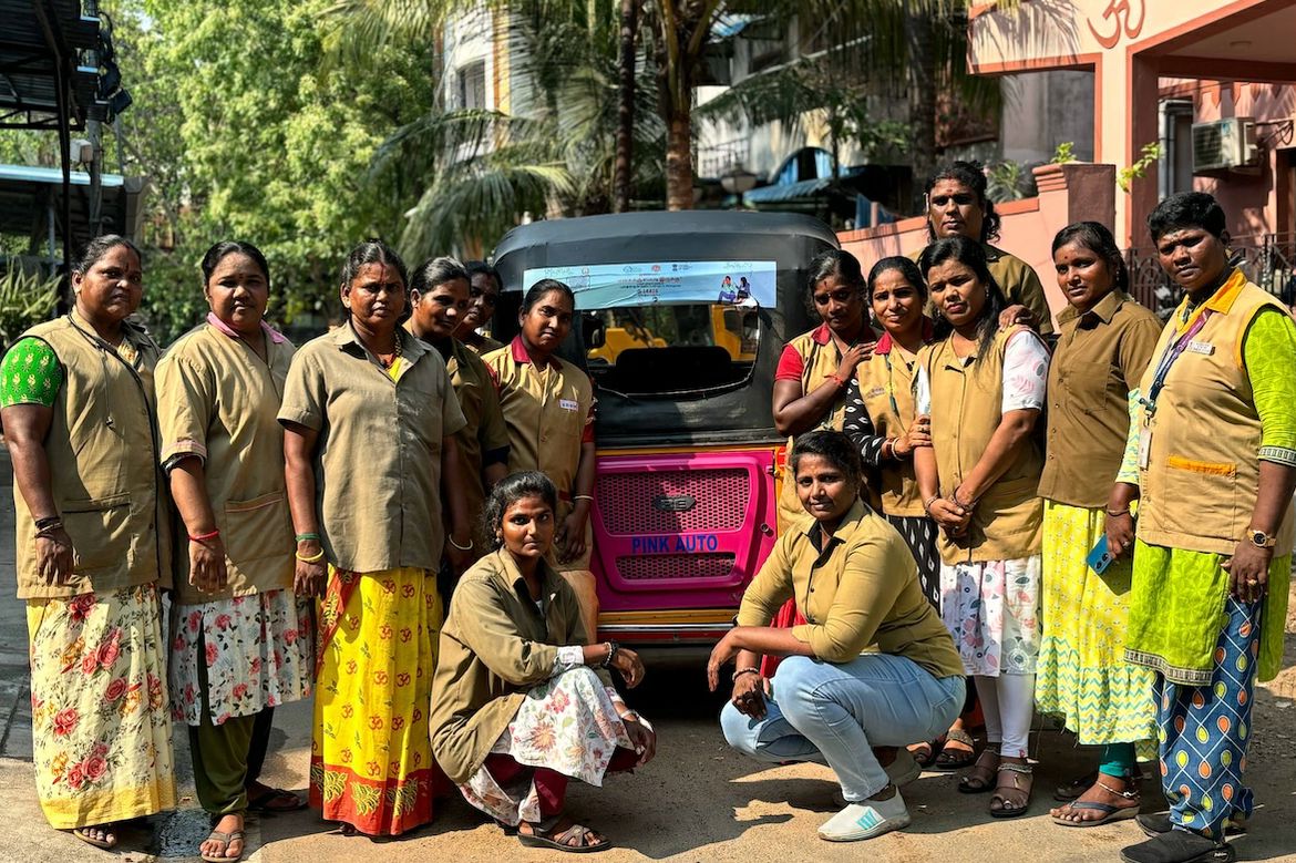 Mohana Sundari poses with members of a women’s empowerment union gathered around a pink auto-rickshaw on a tree-lined street.
