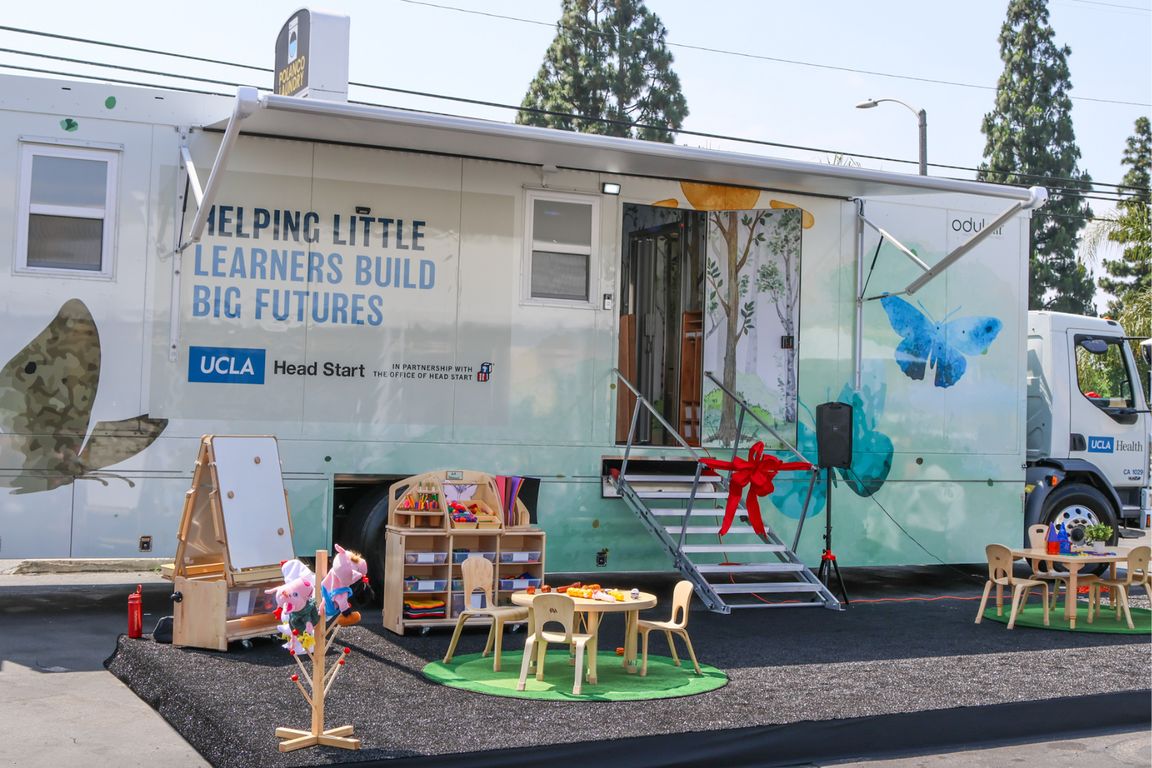 White mobile trailer steps are adorned with tied red ribbon for opening ceremony while gray mat sits underneath two child-size wooden tables small wooden bookcase and white dry-erase board.