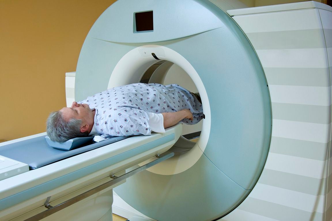 A man with gray hair and a beard lies on his back on a scanner table, wearing a patterned hospital gown, as he is moved into the circular opening of a large MRI or CT scanner in a medical exam room.