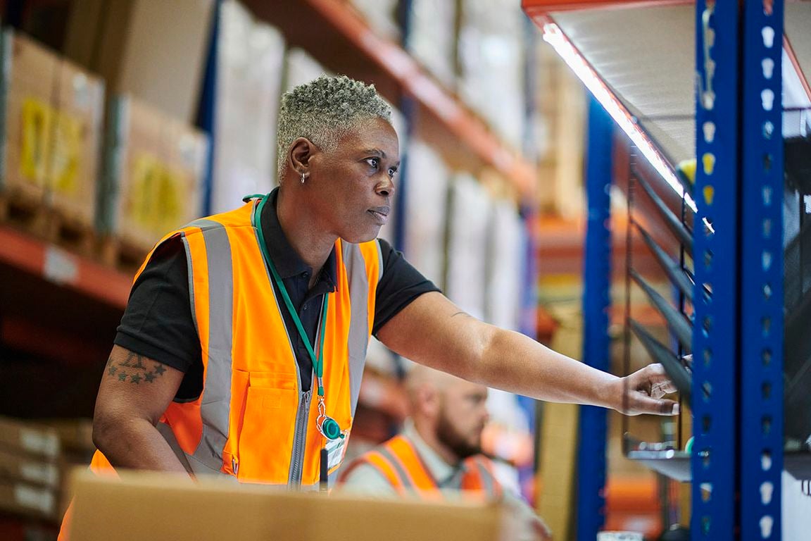 A warehouse worker wearing an orange high-visibility safety vest and green lanyard reaches toward a metal shelving unit stocked with boxes.