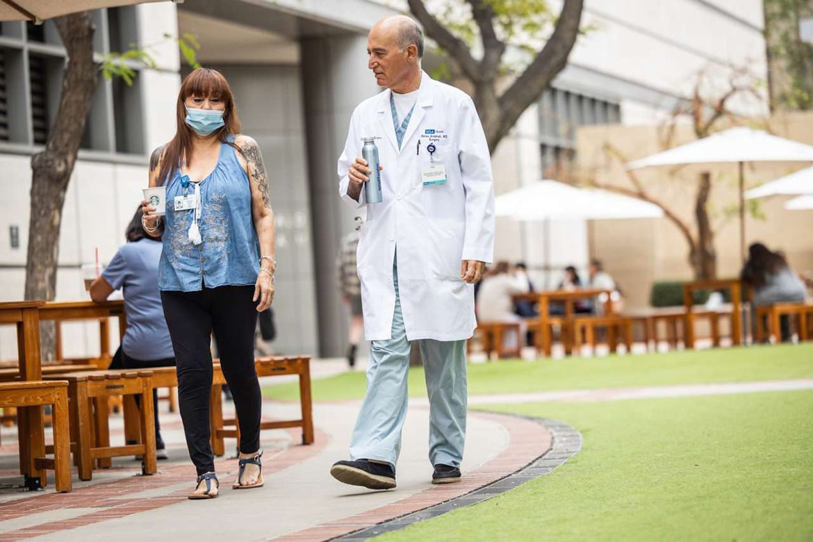 Dr. Abbas Ardehali, wearing a white lab coat over scrubs, walks outdoors with patient Patty Coles, who is wearing a protective mask.