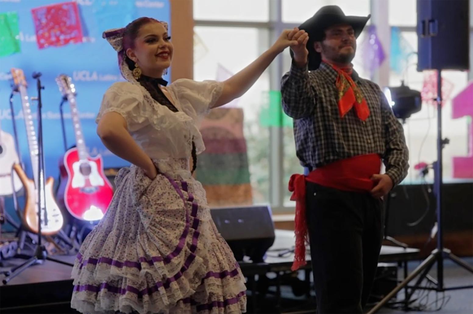 Two dancers in traditional folklórico-style attire perform on an indoor stage, holding hands mid-step. Colorful decorations, guitars, and event equipment are visible in the background.