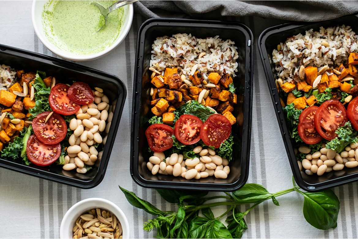 Three meal containers with rice, roasted sweet potatoes, white beans, kale and tomatoes, alongside a bowl of green soup and fresh basil.