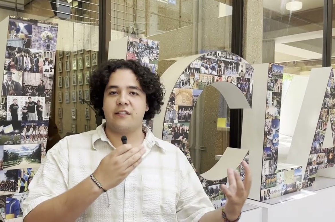 A person holds a microphone in front of a large collage-covered letter display inside a building lobby.