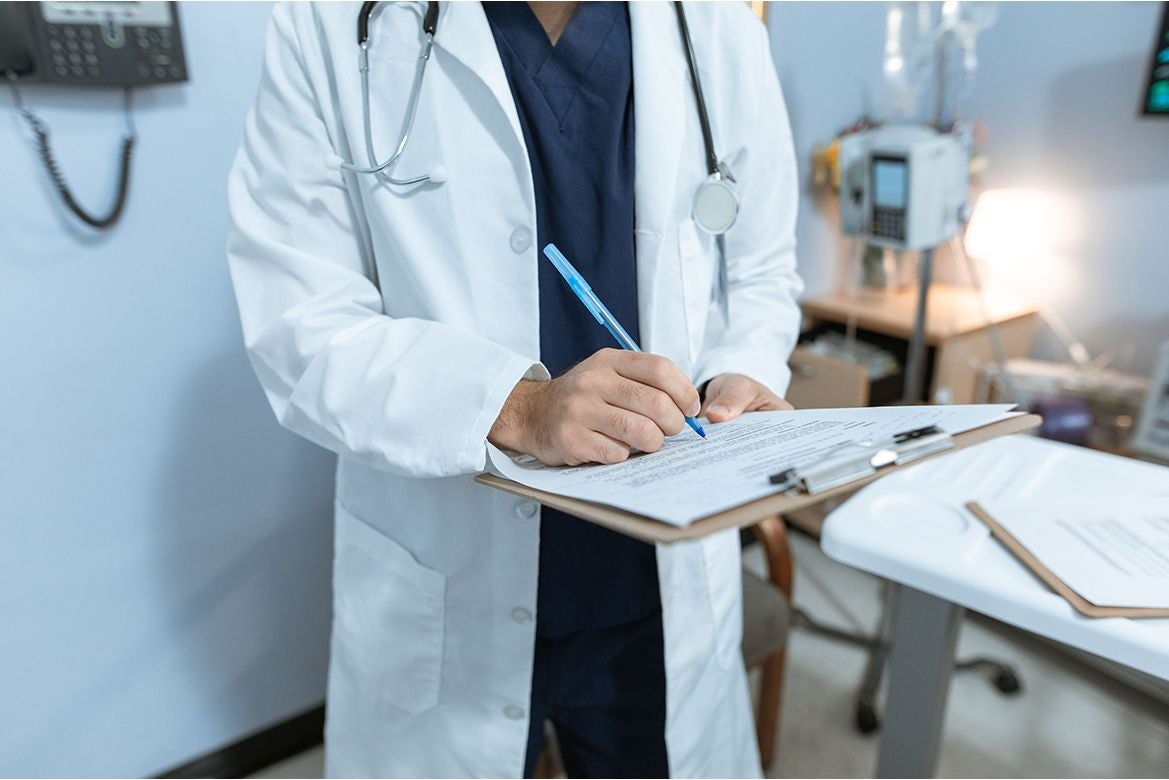Doctor in a white coat writes on a medical clipboard in a clinical office.