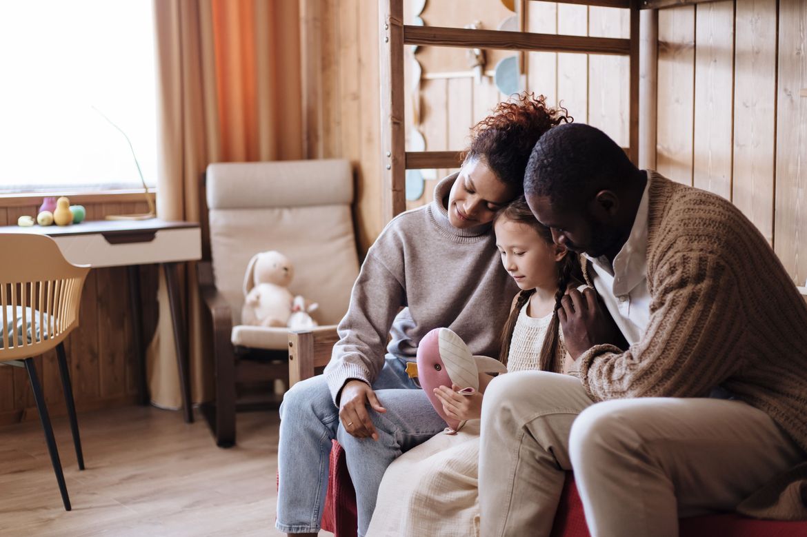 A family sits closely together on a sofa, reading a picture book with a young girl in a cozy wood-paneled room.