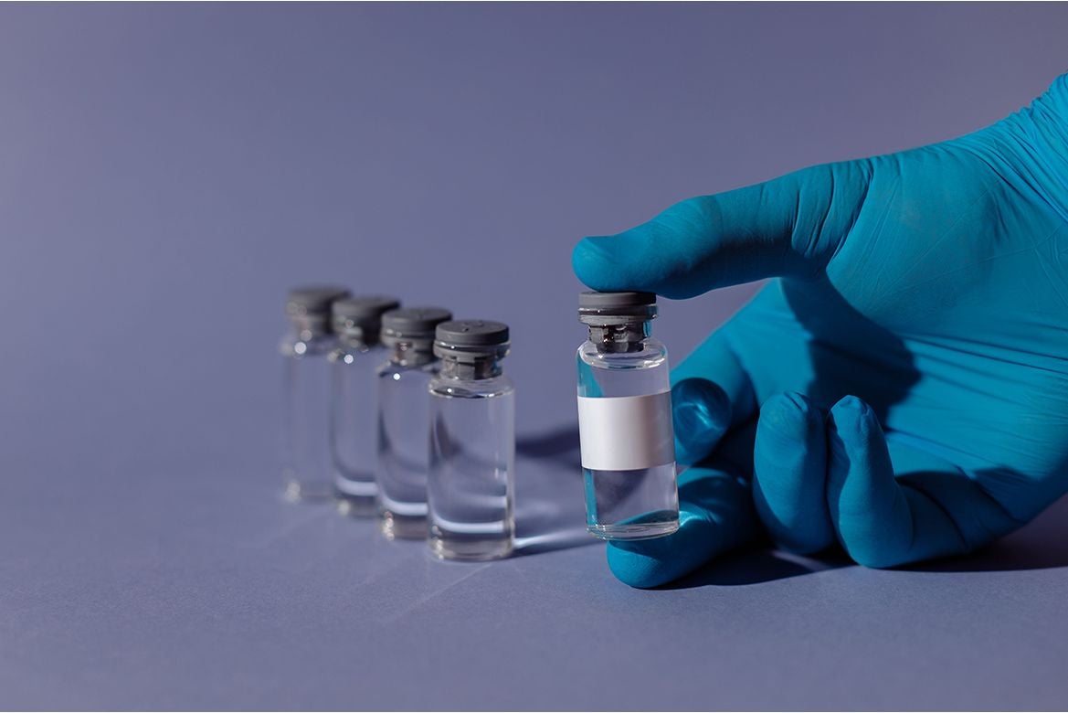 A gloved hand holds a clear medical vial while four additional vials stand in a row on a purple surface.