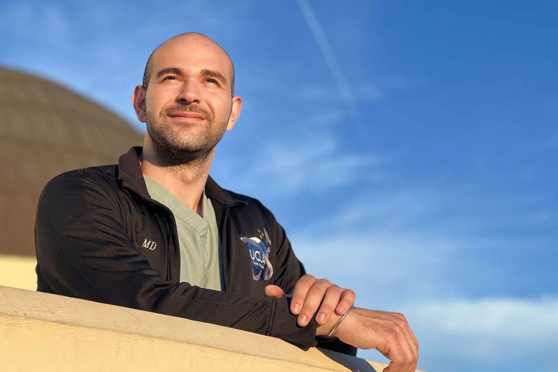 Dr. Haig Aintablian, wearing a green scrub top under a dark zip-up jacket embroidered with "MD" and a UCLA emblem, leans forward on a ledge with arms crossed, smiling and looking up toward the sky.